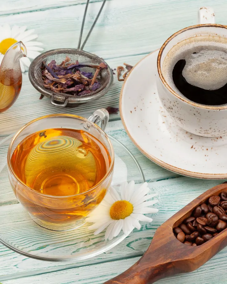 قوری چای شیشه‌ای و فنجان چای کمرنگ کنار فنجان قهوه روی میز فیروزه‌ای