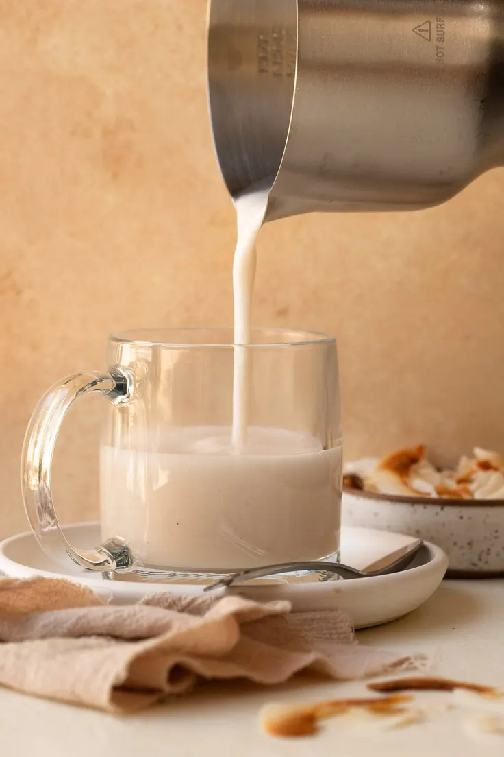 ریختن شیر نارگیل غلیظ از پارچ فلزی در لیوان شیشه‌ای برای قهوه نارگیلی، کنار کاسه پرک نارگیل