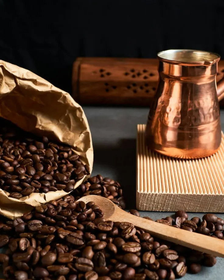 دانه قهوه در پاکت کاغذی کنار بریکی مسی و قاشق چوبی روی میز