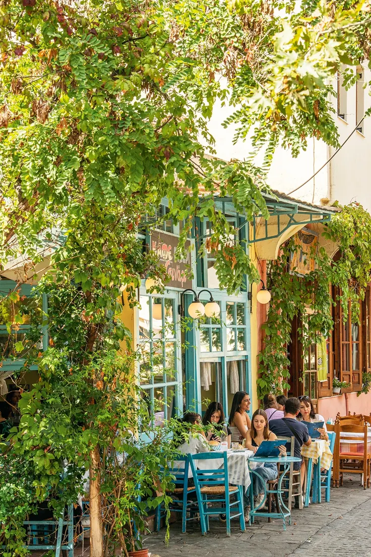 کافه خیابانی یونان با صندلیهای فیروزهای و مشتریهای نشسته زیر درخت
