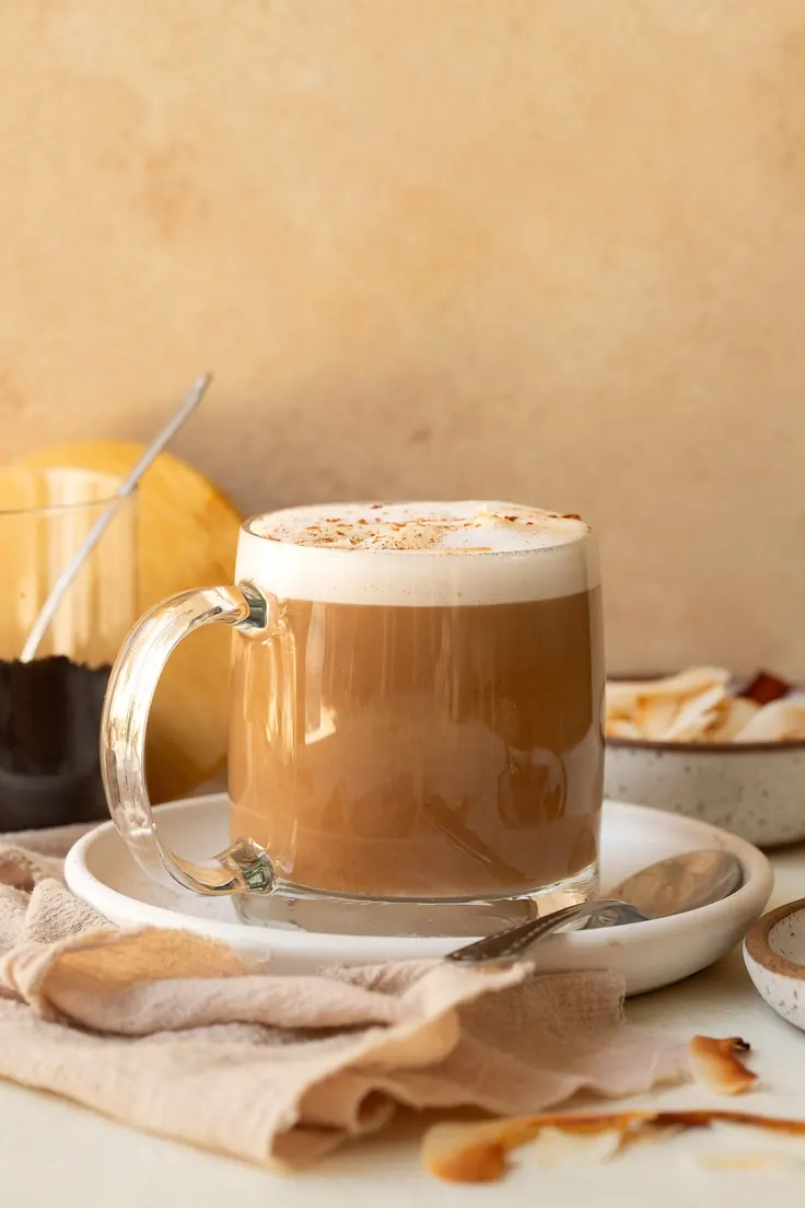قهوه با عسل در لیوان شیشه‌ای با کف شیر و دارچین، کنار شیشه عسل روی میز روشن