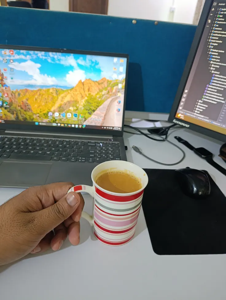 فنجان قهوه شیری روی میز کار واقعی کنار مانیتور و لپ‌تاپ