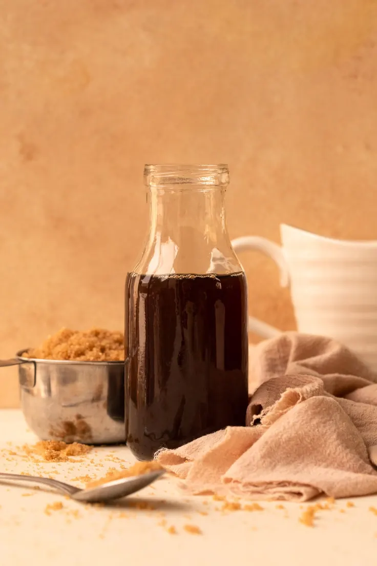 شیشه شربت کارامل خانگی تیره کنار شکر قهوهای و قاشق