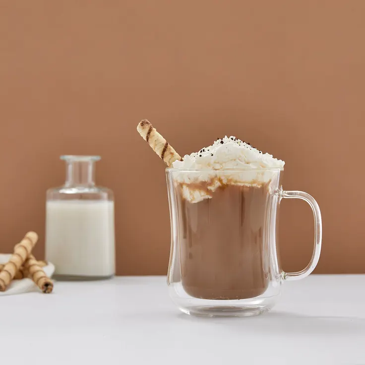 فنجان شیشه‌ای نوشیدنی گرم کرم‌ای با کف شیری روی میز روشن، سرو قهوه گردو خانگی